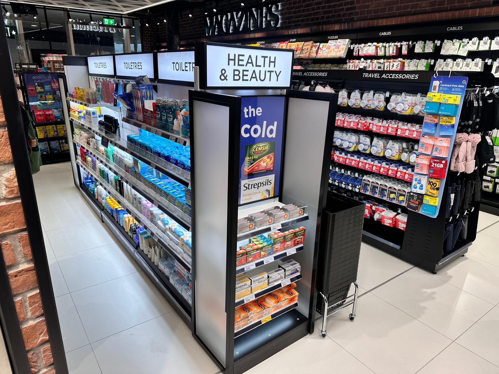 A brightly lit store aisle labeled "Health & Beauty" displays toiletries, cold medicine, and personal care products. Nearby shelves hold travel accessories, with a shopping basket in the foreground.