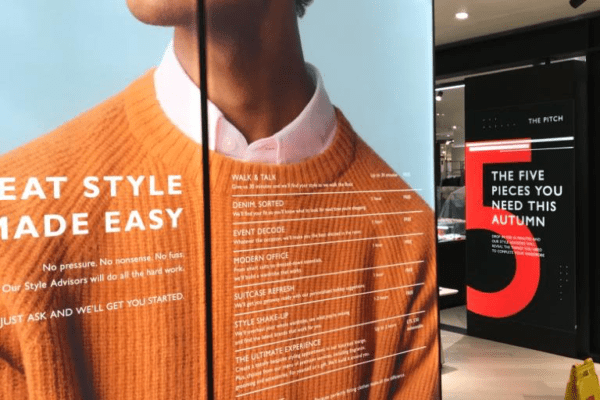 An image of a retail store display featuring large, LED backlit graphics. The left panel shows a person wearing an orange sweater with text about style advice. The right panel has a red '5' with the heading "THE FIVE PIECES YOU NEED THIS AUTUMN." A caution sign is on the floor.