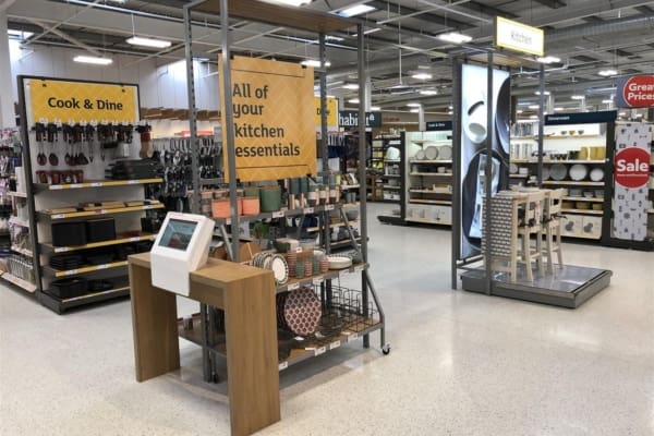 A spacious store section with kitchen essentials, featuring a vibrant LED lightbox reading "All of your kitchen essentials." Shelves stocked with cookware, tableware, and kitchen tools are visible. A display table in the center showcases various items. Eye-catching backlit graphics indicate sales and price reductions.