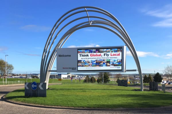 A large welcome sign at the entrance of Aberdeen International Airport features an arched metal structure with LED architectural illumination. An LED screen displays the message, "Think Global, Fly Local" along with promotional travel information. The background includes a blue sky and green grass.