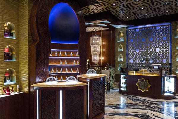 A luxurious, dimly lit fragrance shop. Ornate wooden shelves, enhanced with LED shelf lighting, display various perfume bottles. Intricate decorations adorn the walls and ceiling. Two central counters are illuminated from below, and the marble floor adds a touch of elegance to the atmosphere.