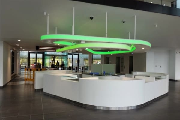 A modern reception area with a large, white, curved desk and LED ceiling illumination, featuring a futuristic green light fixture hanging above. The space has a sleek design with glass partitions and an open layout. In the background, there are large windows letting in natural light.