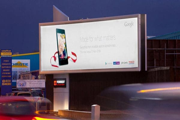 A highway billboard at dusk, featuring LED backlit graphics, displays an advertisement for Google's Nexus devices alongside logos from Tesco and Carphone Warehouse. Cars pass by in the foreground, while buildings with bright signage illuminate the background, epitomizing out of home advertising.