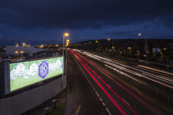 Nighttime highway scene with light trails from fast-moving vehicles in both directions. To the left, a large illuminated billboard featuring LED backlit graphics displays a 20th-anniversary advertisement. In the distance, city lights are visible under a dark, cloudy sky.