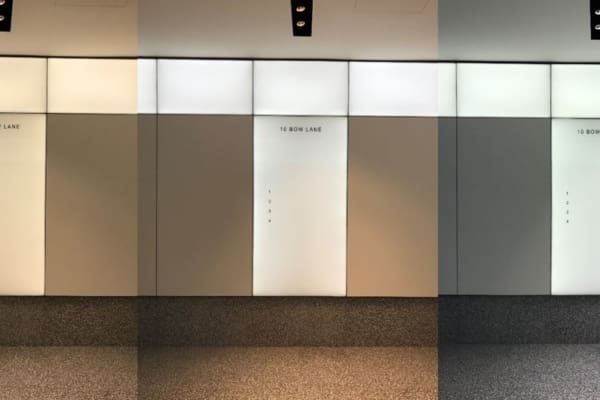 A modern indoor wall with a textured, speckled floor. The wall features muted grey panels, with the middle panel illuminated by LED light panels and displaying the address "10 Bow Lane" in small, black text. The left and right panels are in shadow, creating a symmetrical look enhanced by subtle colour temperature adjustments.