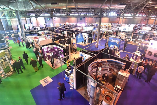 A bustling trade show floor with numerous booths displaying various products and services. Attendees are walking around, engaging with exhibitors, and exploring the different displays. The floor is covered in sections of green and blue carpet, and the venue is well-lit.