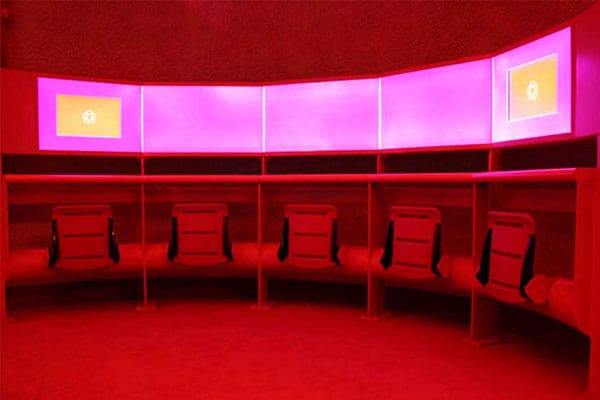 A room illuminated by red LED lighting features a curved wall adorned with multiple rectangular light panels displaying a red flag with a yellow emblem. Beneath the screens, there are seven office chairs arranged in a semicircle.