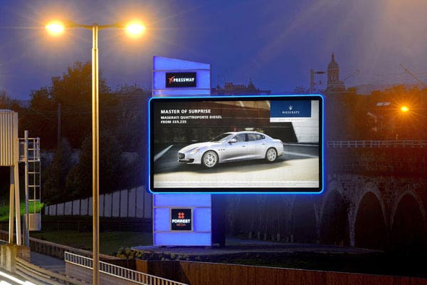 A large digital billboard at night, illuminated with crisp LED lighting, displays an advertisement for a Maserati Quattroporte Diesel with the tagline "Master of Surprise." Blue accents light up the sign, while a brightly lit street lamp stands in the foreground. Trees and a building fill the background.