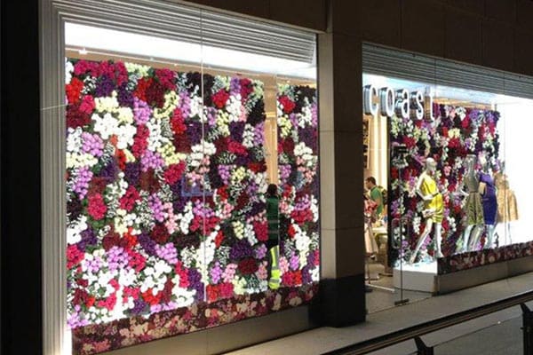 A store display window adorned with a wall of colorful flowers in various shades of pink, purple, white, and red is elegantly lit by LED window lighting. Mannequins dressed in fashionable clothing stand in front of the floral backdrop, with the store's name partially visible on the right side.