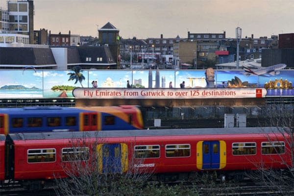 A train passes in the foreground, with an Emirates LED out of house advertising billboard in the background. The advertisement displays various exotic destinations and reads, "Fly Emirates from Gatwick to over 125 destinations." Urban buildings and rooftops are visible behind the billboard.