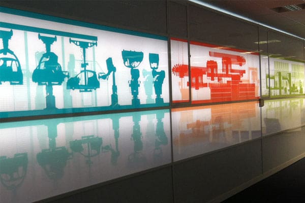 An illuminated, futuristic wall display featuring abstract silhouettes in shades of cyan, red, and green. The LED light panel illumination enhances images resembling various mechanical and robotic components, mounted on a reflective white surface that extends along a corridor or hallway.