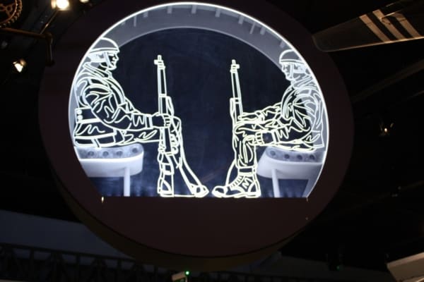 A circular display features an illuminated art piece of two soldiers sitting face-to-face and holding rifles, enhanced by LED glass art illumination. Set against a dark background, the artwork appears suspended in mid-air, with the soldiers sketched in white lines and details.