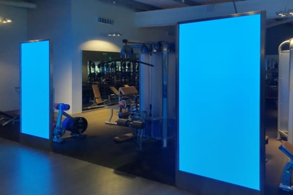 A gym interior with various workout equipment, including weight machines and free weights. Prominent large, blue LED light sheet panels are set up on either side of the gym's entrance area, casting a blue hue over the space. The room is dimly lit, highlighting its unique architectural features.