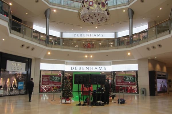 A two-story shopping mall featuring an open central atrium. The ground floor has a Christmas tree adorned with decorations and a setup for a performance. Both the upper and lower levels boast Debenhams stores, with New Look visible on the lower level, highlighted by vibrant brand illumination from LED light sheet panels.