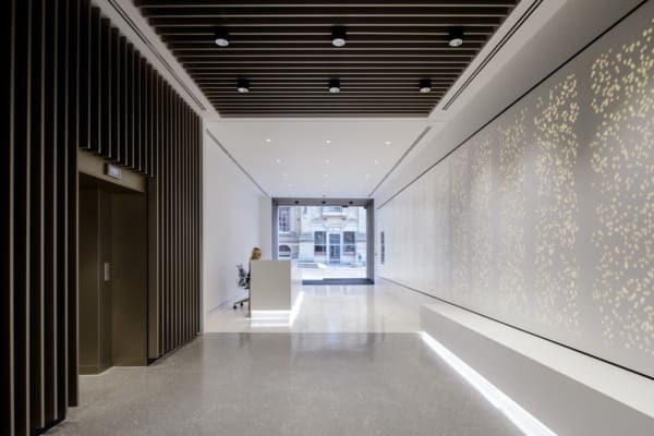 A modern building lobby featuring a minimalist design with sleek lines and neutral tones. On the left is a wooden slat wall with wall illumination, leading to an elevator. A receptionist sits at a white desk near the entrance, which offers a view of the urban street outside.