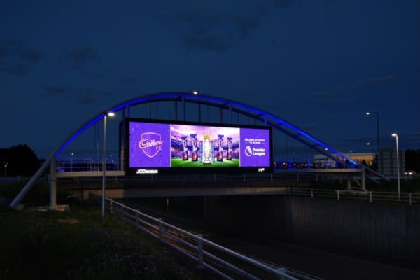 A large, illuminated digital billboard on a bridge displays an advertisement for the Premier League with the slogan "Celebrating 30 years of the Premier League." It is nighttime, and the surroundings are dimly lit, accentuated by LED architectural illumination that highlights the bright colors on the billboard.