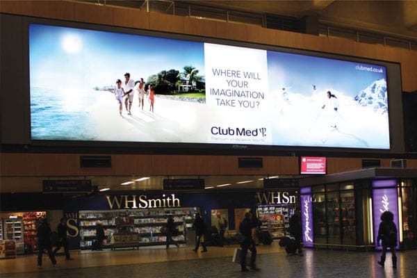 A large illuminated billboard with LED backlit graphics for Club Med is displayed indoors above a WHSmith store. The advert features an image of people walking on a beach with the text, "Where will your imagination take you?" The setting appears to be a busy public space.