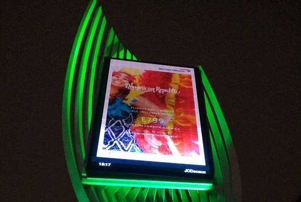 An illuminated digital billboard with a vibrant advertisement for British Airways promoting flights and holidays to the Dominican Republic. The sign highlights a special offer of £799, including all-inclusive resorts. The surrounding structure glows with a colour-changing LED lighting system.