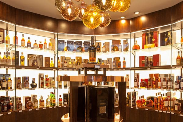 A well-lit liquor store display with numerous bottles of various spirits arranged in a semi-circular glass case. Shelf lighting and LED light panels enhance the visibility of each bottle. Golden spherical pendant lights hang from the ceiling, adding a luxurious touch to the setting. A central pedestal showcases a featured bottle.