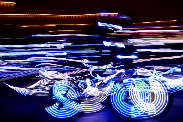 A cyclist adorned with blue LED linear lighting rides in the dark. The motion creates dynamic, swirling patterns of light, transforming the scene into a vibrant, abstract display of streaks and trails against a dark background. The surroundings are illuminated with various light trails.