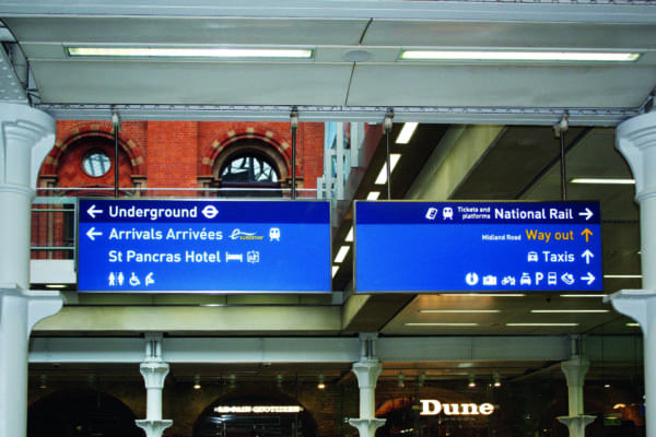 A signboard at St Pancras International Station using an LED wayfinder directs passengers to the Underground, Arrivals, St Pancras Hotel, National Rail, Taxis, and the Way out. The blue signs display icons for transportation and facilities against a visible red brick wall in the background.