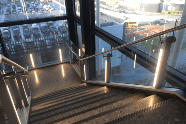 A modern stairwell with metal railings and LED illuminated handrails leads downwards. Large windows on the sides provide a view of a sunlit parking lot and city street outside. The stairwell is well-lit with natural light streaming in.