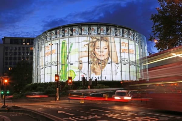 A large building at dusk with a curved glass exterior displaying a brightly illuminated LED out of home advertising for Rimmel featuring a woman's face. The streets around the building have light trails from passing vehicles, and buildings and trees are visible in the background.