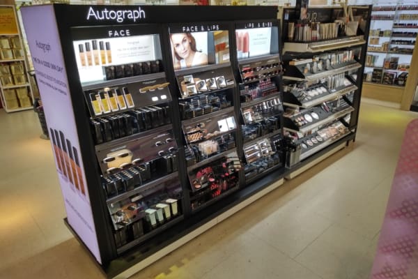 A store display featuring various makeup products from the brand "Autograph" with product illumination provided by an LED light panel. It includes sections for face products, face and lip items, and an assortment of cosmetics like foundations, concealers, lipsticks, and powders, arranged neatly on shelves.