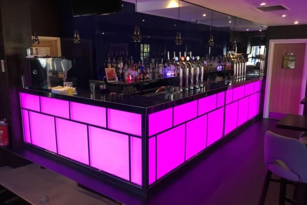 A modern bar with a vibrant purple-lit counter featuring LED light sheet panels, various beer taps, and shelves stocked with bottles and glasses. The illuminated bar area boasts a sleek, contemporary design with pendant lights, a coffee machine on the counter, and seating areas in the foreground and background.