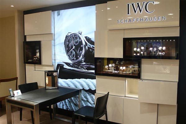 A sleek interior of a watch boutique displaying luxury timepieces from IWC Schaffhausen. The foreground features a modern table and two chairs, while the background showcases illuminated watch displays and a large promotional poster with LED backlit graphics highlighting a black watch model.