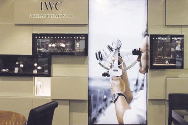 A display showcases luxury watches at an IWC Schaffhausen store. The centerpiece features an LED backlit graphic of a person using a sextant, with the person wearing an IWC watch. To the left and right of the image are glass display cases holding various watch models.