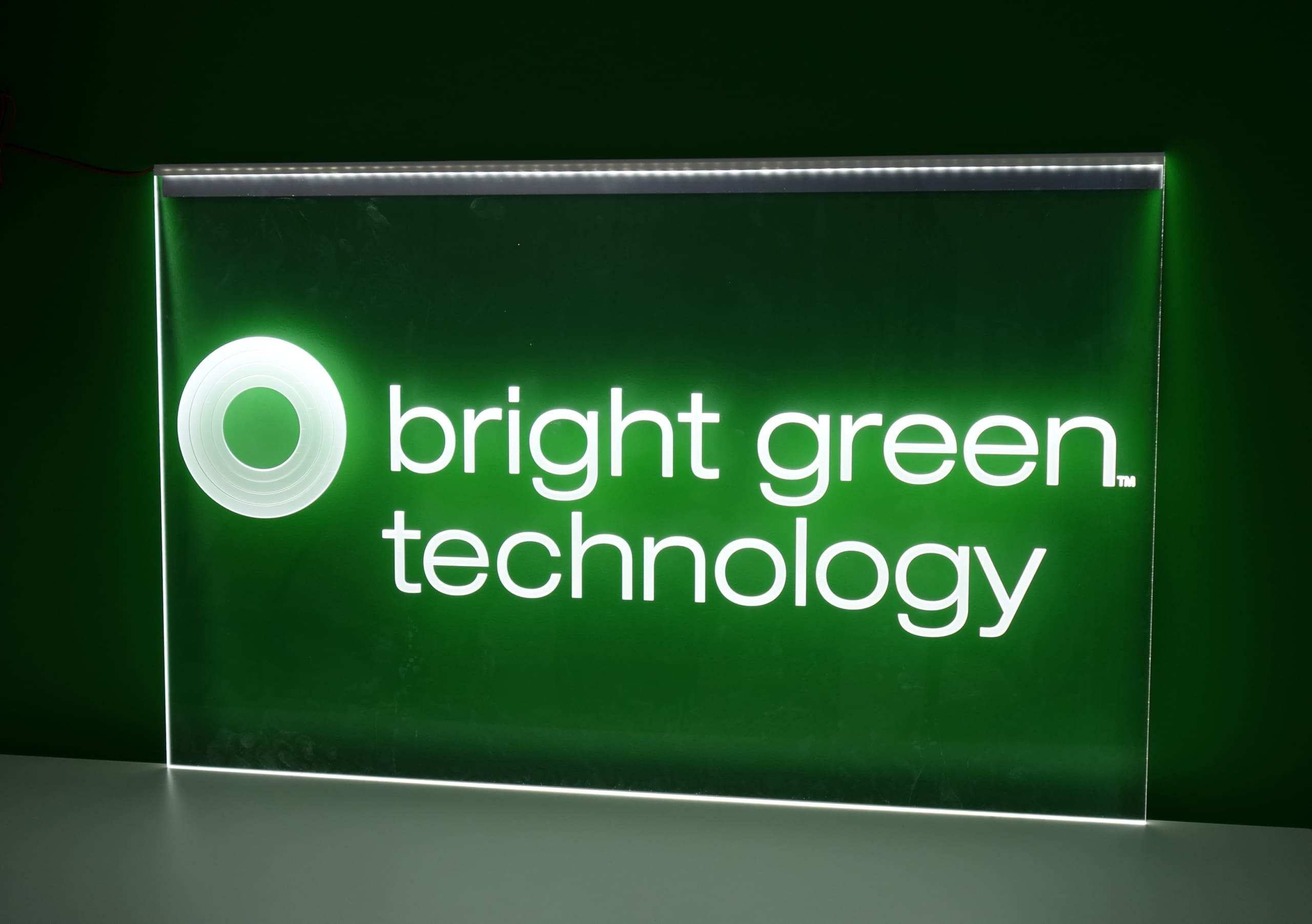 A green and white illuminated sign reads "bright green technology" with a circular logo on the left. The text and logo are brightly lit against a darker green background.