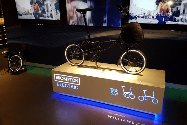A Brompton Electric folding bike is displayed on a raised platform with a backlit edge. The background features large screens showcasing the bike in different settings. The display includes icons and text highlighting the bike's features and design collaboration, enhancing brand illumination.