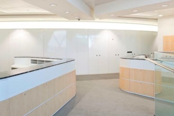 A modern, well-lit interior space with a sleek design featuring curved reception counters, light wooden panels, and white walls. LED ceiling illumination enhances the area, while a minimalistic glass railing contributes to the clean and spacious feel.