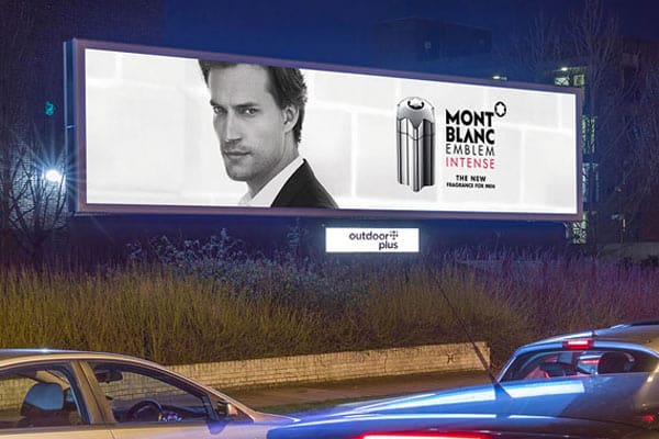 A large outdoor billboard at night displays an advertisement for Mont Blanc Emblem Intense, a fragrance for men. The billboard, enhanced with LED backlit graphics, features a black-and-white photo of a man in a suit next to an image of the product. Cars are seen driving past this out-of-house advertising setup.