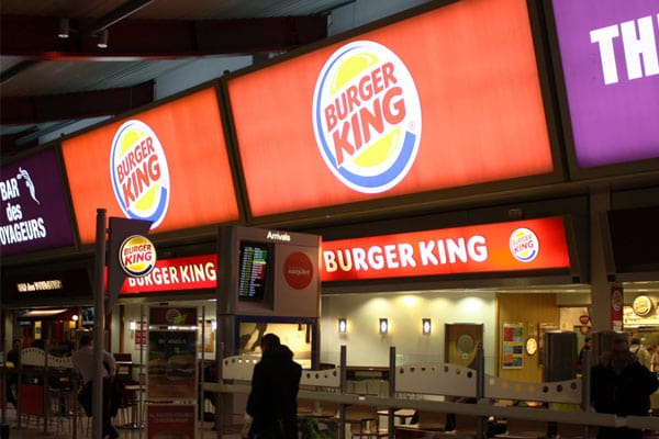 A Burger King restaurant is illuminated with bright LED backlit graphics at night. The signs display the Burger King logo and name in large, bold letters. There are a few people standing near the entrance and an arrivals board visible in the background.
