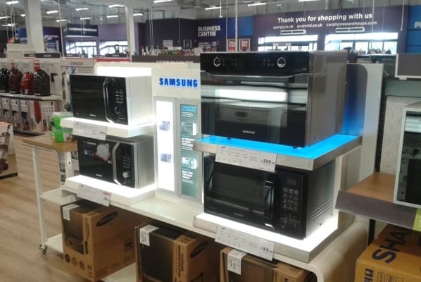 An electronics store display showcases various Samsung kitchen appliances, including microwaves and ovens, on a shelf. The appliances are illuminated with bright lights, likely from LED light sheet panels, and their prices are visible. Boxes are stacked below the shelf, with other store sections in the background.
