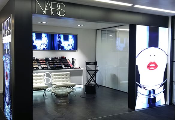 A modern NARS cosmetics store featuring a sleek black and white design. Inside are displays of makeup products, two large video screens playing content, a glass table with white chairs, and an illuminated image of lips with red lipstick on the front right wall, enhanced by LED backlit graphics.