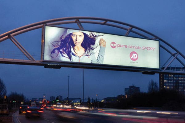 A large double-sided LED billboard over a busy highway at dusk shows a woman in athletic wear with long hair flowing, captioned with "pure simple SPORT" and "JD SPORTS" alongside their respective logos. Vehicles drive below the sign, and the city skyline is visible in the background.
