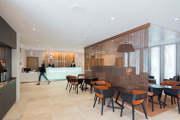 A modern hospital lobby features a sleek reception desk with an illuminated sign reading "Spire Cambridge Lea Hospital." The area, bathed in natural light, includes a comfortable seating space with black and orange chairs around small tables, and a glass partition with a textured design.