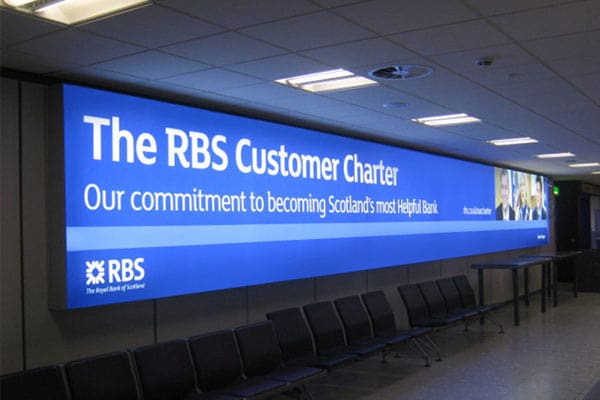 Large blue advertisement on a wall in an indoor setting, displaying "The RBS Customer Charter" with the tagline "Our commitment to becoming Scotland's most Helpful Bank." Enhanced by LED back lit graphics, the RBS logo and "The Royal Bank of Scotland" are also visible along the bottom.