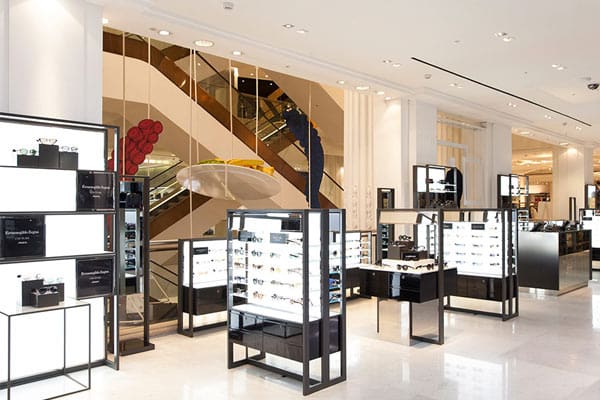A modern, well-lit retail store with glass display cases showcasing eyewear. The background features a sleek escalator adorned with red and blue sculptures, leading to an upper level. Enhanced by LED light panels, the store boasts a clean, minimalist design with white walls and shiny floors.