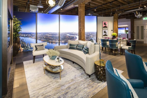 A modern living room features a curved white sofa with accent pillows, a round coffee table, and an area rug. Behind the sofa, illuminated LED graphics enhance the large panoramic cityscape. There are two teal armchairs, a wooden ceiling with exposed ducts, and a nearby dining area with a table and chairs.