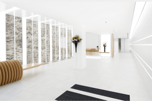 A modern, minimalist interior with white walls and flooring. A large mirror, floral arrangements, and a unique, round wooden bench decorate the space. Vertical white slats partially cover a textured wall on the left, while sleek horizontal lighting and an LED light sheet panel accent the right wall.