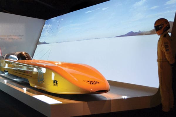 An aerodynamic yellow race car is displayed on a platform under bright illumination, with LED backlit graphics in the background showing a shiny, reflective surface and a clear sky. Next to the car, a person in a yellow racing suit and helmet stands to the side.