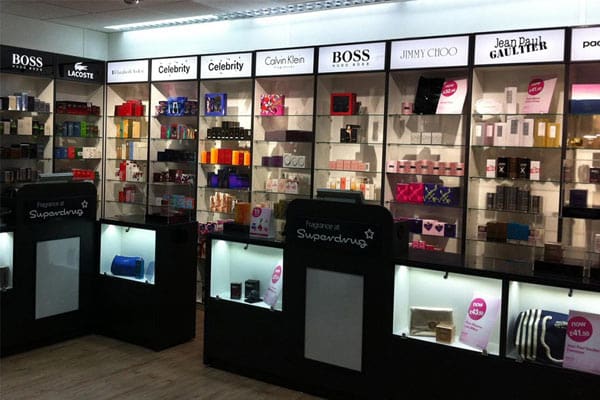 A well-lit retail display featuring various high-end fragrances and beauty products from brands like Boss, Lacoste, Calvin Klein, Jimmy Choo, and Jean Paul Gaultier. Shelves with LED shelf-lighting are neatly organized with backlit brand logos and product boxes arranged methodically.