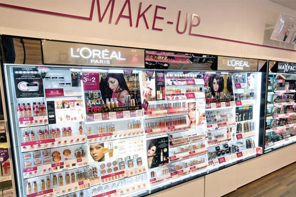 A well-lit store display showcases a wide variety of L'Oréal Paris makeup products, including foundations, lipsticks, mascaras, and eyeshadows. The shelves are neatly organized under an LED light panel, and promotional posters highlight specific items and offers. The word "MAKE-UP" is displayed above.