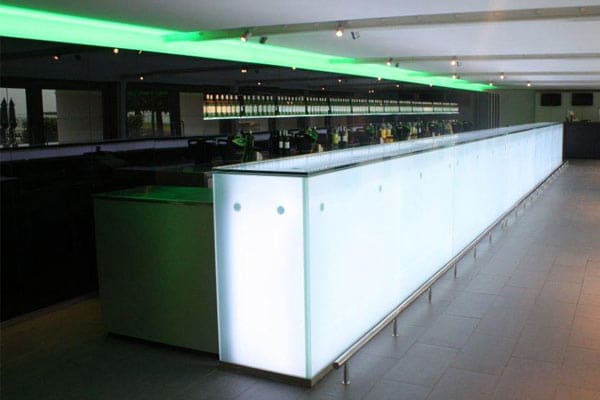 A modern bar with a sleek, illuminated counter top features a long, white light-up counter with a green-lit accent above, enhanced by an LED light panel for backlit bars. Various bottles and glasses are arranged neatly behind the bar. The floor is tiled in gray, cementing the contemporary ambiance.