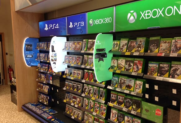 A retail display featuring shelves stocked with video game titles for PlayStation 4, PlayStation 3, Xbox 360, and Xbox One. Large, LED brand illumination signs for each console hover above the respective sections, showcasing a variety of game options.
