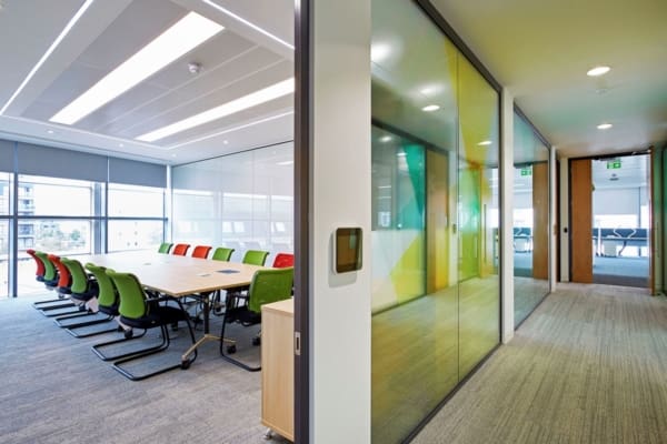 A modern office meeting room with a long wooden table surrounded by colorful chairs. The room features large windows with shades, glass partition walls, and a hallway leading to additional office spaces. Enhanced with ceiling illumination from LED light sheet panels, the environment is well-lit and neatly organized.